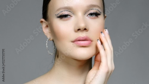 Female with unusual rhinestones makeup and earrings in the ears. Hand near face.