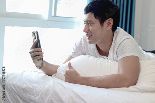 Asian man using smartphone in bed after wakening up in the morning in his bedroom. A man checking his mobile phone while lying down on comfortable bed. Sleepy man holding smartphone in bed at home.