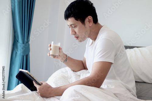 Asian man using tablet while drinking almond milk in bed after waking up in the morning in bedroom. Man working on his tablet during lying down on comfortable bed. Sleepy man surfing internet in bed.