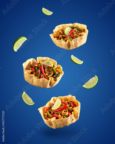 Three tortilla bowls with fajitas with beef, cheese, corn and lettuce and lime wedges levitate on blue background