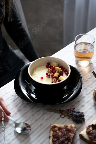 Bowl of semolina porridge with walnuts, red currants and apples on a black plate. Woman’s hand with spoon, tea and toast on cozy morning breakfast table