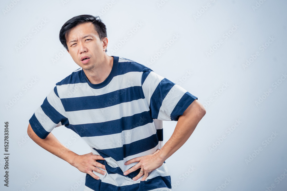 Fototapeta premium Asian man in striped shirt showing discomfort and fatigue, leaning forward with hands on waist and pained expression, standing against plain studio background