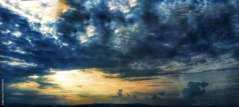 Obraz premium A dramatic cloudy sky with dark, swirling clouds and sunlight piercing through, casting striking beams of light that create a powerful and captivating atmosphere. 