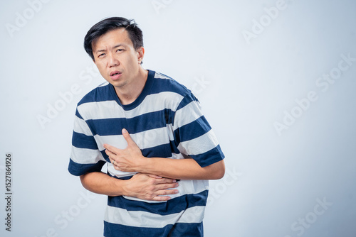 Asian man in striped shirt showing pain and discomfort while holding his chest and stomach, possibly indicating heartburn or illness, standing against plain studio background