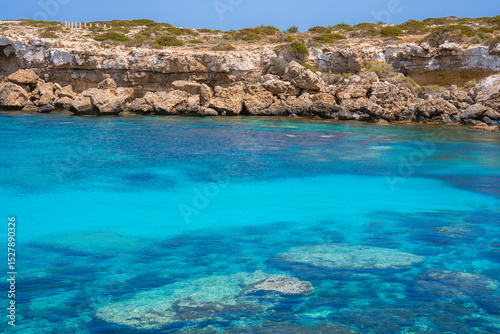 Crystal clear turquoise waters with visible underwater rocks meet rugged light colored cliffs topped with sparse vegetation along Cyprus' coastline.