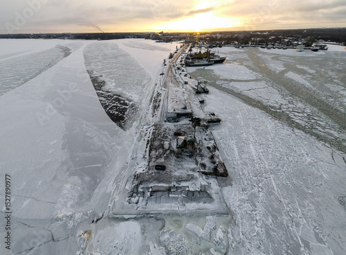 An aerial view of a snow and ice covered port with several ships docked, set against a dramatic winter sunset over the frozen bay