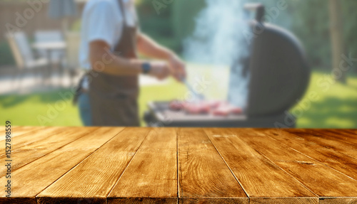 Empty wooden picnic table with copy space and blurred garden background of a person grilling on the grill and having summer leisure time concept.