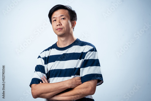 Asian man in navy and white striped t-shirt crossing arms while looking confidently at the camera, posing against plain light background, conveying attitude, confidence, and modern style