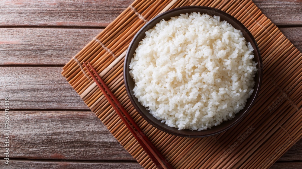 A top-down view of a bowl of hot, fluffy rice with steam rising, placed on a bamboo mat with chopsticks resting beside it for an authentic and traditional look. A top-down view of a bowl of hot, fluffy rice with steam rising, placed on a bamboo mat with chopsticks resting beside it for an authentic and traditional look.