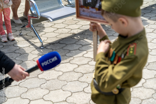 Young boy dressed in military uniform holding a photo while reporters interview at a commemorative event in springtime