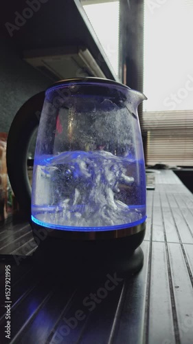 A close-up shot captures the modern and energetic mood of an electric glass kettle, illuminated by a blue LED light, as water vigorously boils inside, showing the efficiency of modern kitchen