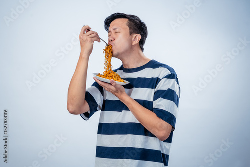 Asian man in striped shirt eating a mouthful of spicy instant noodles from a bowl with a fork, eyes wide open, captured against a plain background