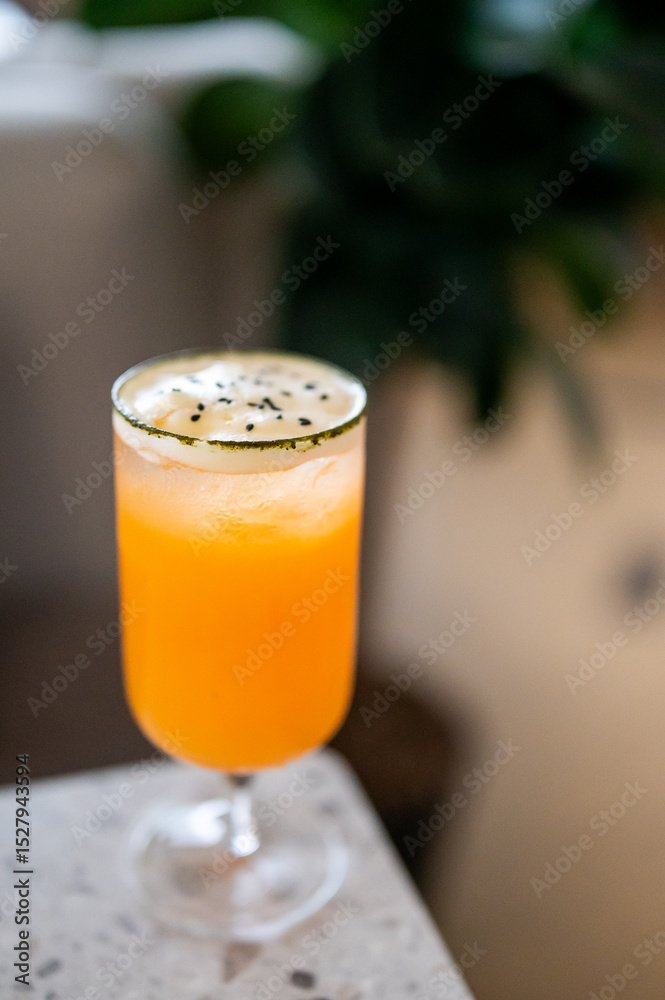 A vibrant orange cocktail served in a tall glass with frothy white foam, garnished with black sesame seeds. Placed on marble surface with a blurred background, highlighting its refreshing presentation A vibrant orange cocktail served in a tall glass with frothy white foam, garnished with black sesame seeds. Placed on marble surface with a blurred background, highlighting its refreshing presentation