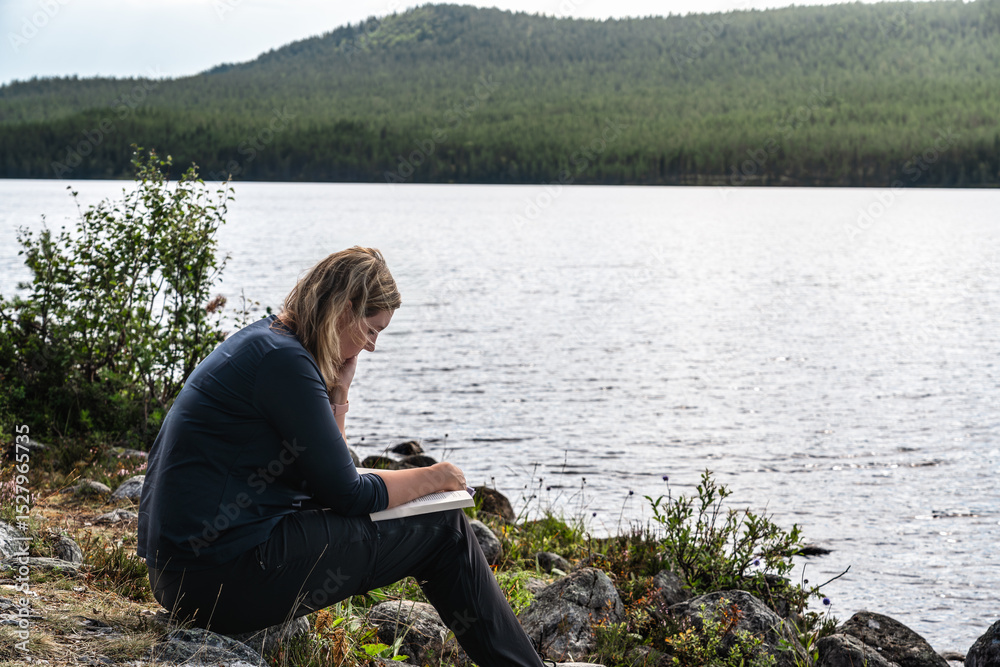 Fototapeta premium A woman is reading a book while sitting at the shore of a lake on a bright summer day in Sweden. She is enjoying the peace and quiet of nature. 