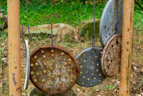 Rusty perforated metal lids hanging on wires between posts, serving as improvised garden chimes. Greenery and stones are visible in the background.