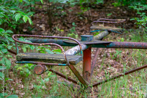 old, rusty playground carousel with metal bars and wooden seats, located in an overgrown area surrounded by grass and bushes. A faded remnant of childhood play in an abandoned place.
