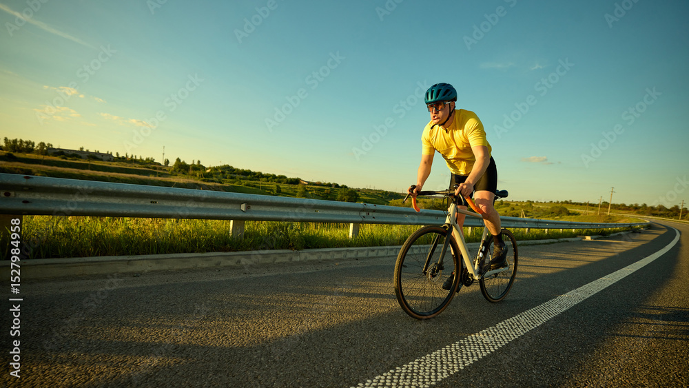 Fototapeta premium Man rides hard on wide road through nature terrain with intense expression against hills and soft sunset on background. Banner. Concept of sport, cycling, healthy lifestyle, endurance, nature.