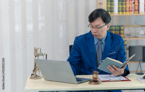 Asian male lawyer working in office with laptop and scales, judge's gavel, reading law book in office for lawyer consulting concept