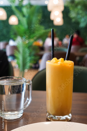 A vibrant mango smoothie in a tall glass with straws, placed on a wooden table, set against a blurred cafe background.