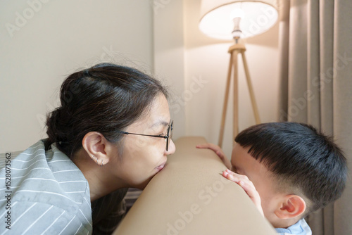 Asian mother and Little child boy playfully playing peekaboo game over couch in cozy room