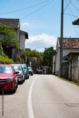 Fototapeta Naklejka Na Ścianę i Meble -  City street features a line of parked cars under a clear blue sky. Traditional stone houses with tiled roofs flank the roadway, creating a serene and picturesque scene.