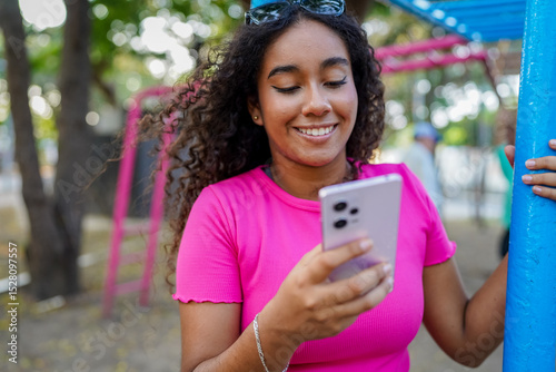 Young woman using mobile phone in a park with playground in the background
