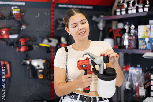 Woman examines handheld wireless portable spray gun, airbrush in tools department of construction store. Girl consider house or apartment renovation redecoration. Interior design, styling concept.