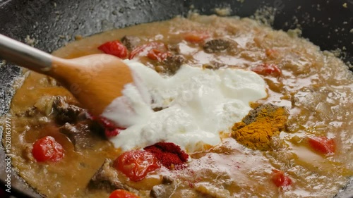 A wooden spatula stirs natural yogurt into a delicious beef stew. The meat is simmering in a creamy sauce with fresh tomatoes and herbs. A closeup of a homemade gourmet meal preparation