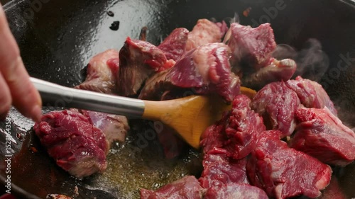 A kitchen spatula stirs raw beef pieces cooking in hot bubbling oil in a black frying pan. Closeup of searing meat a culinary process for preparing a delicious protein rich dinner
