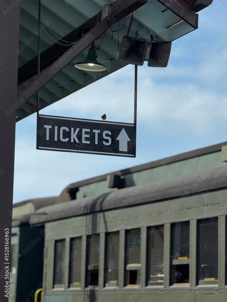 Obraz premium A hanging ticket sign with an arrow, at a train station. A passenger train with windows is visible in the background. A small songbird is perched on the black sign with white letters.