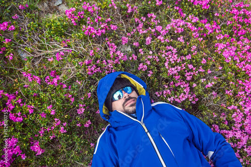 Hiker relaxing in colorful wildflower meadow, surrounded by bright pink blooms while wearing protective outdoor gear and mirrored sunglasse