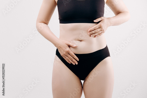 Woman in black underwear is holding her stomach, expressing discomfort and pain, with a neutral background highlighting her emotional state