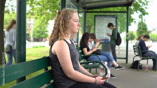 Teen Girl Sitting Still at Bus Stop While Others Scroll Phones