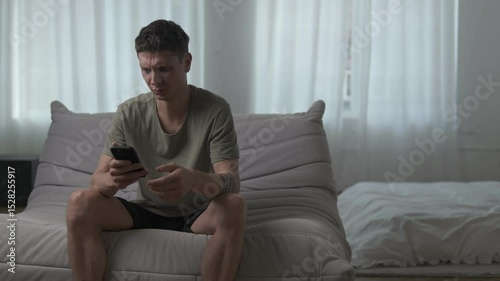 Young man reacts with disgust to something on his phone while sitting on a sofa in a modern apartment.