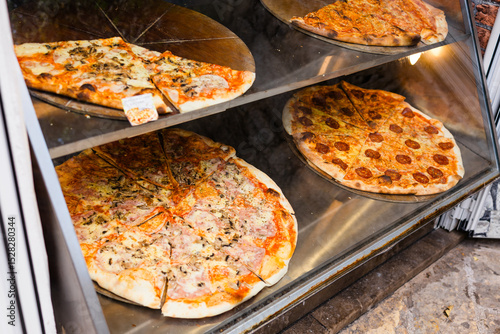 Various pizzas on display in a glass case at a street pizzeria. Fast food, appetite, and casual urban dining.