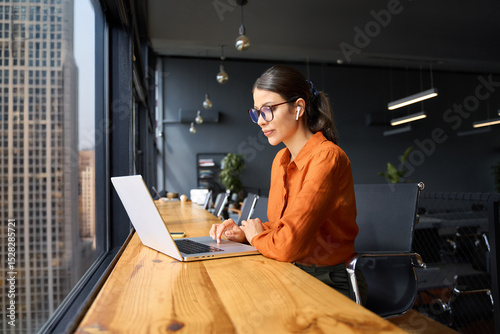 Busy latin hispanic young business woman working on laptop computer sitting at office desk with city view. Indian entrepreneur manager businesswoman using pc for communication, learning at workplace photo