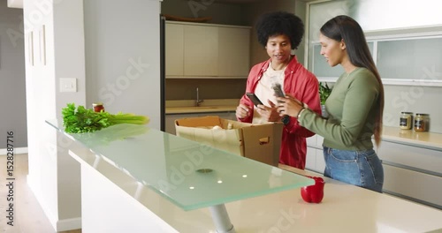 African American and Indian couple unpacking grocery box at counter scanning phone checking pepper