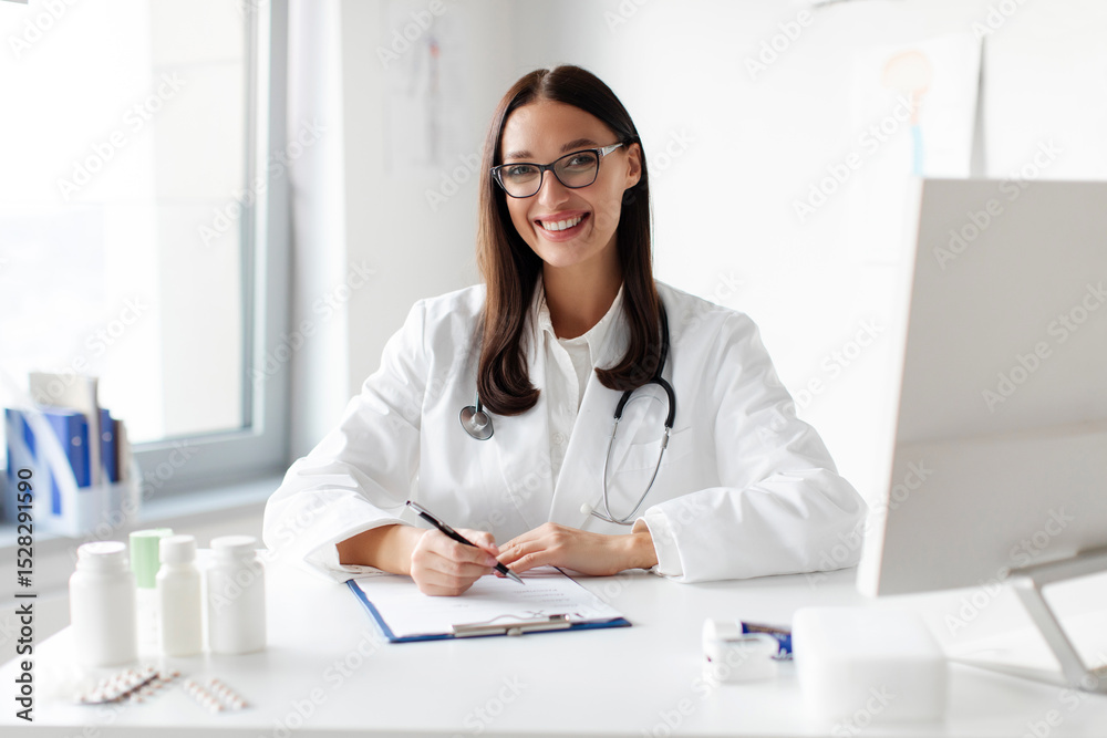 Happy friendly young lady wearing white medical coat with stethoscope posing at workplace at clinic, smiling at camera. Healthcare, medical workers concept Happy friendly young lady wearing white medical coat with stethoscope posing at workplace at clinic, smiling at camera. Healthcare, medical workers concept