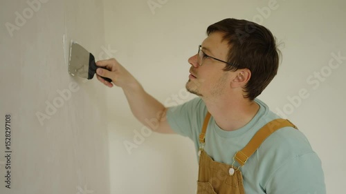 Professional house painter wearing glasses and brown overalls, filling cracks in a white wall using a putty knife and plaster before starting to paint