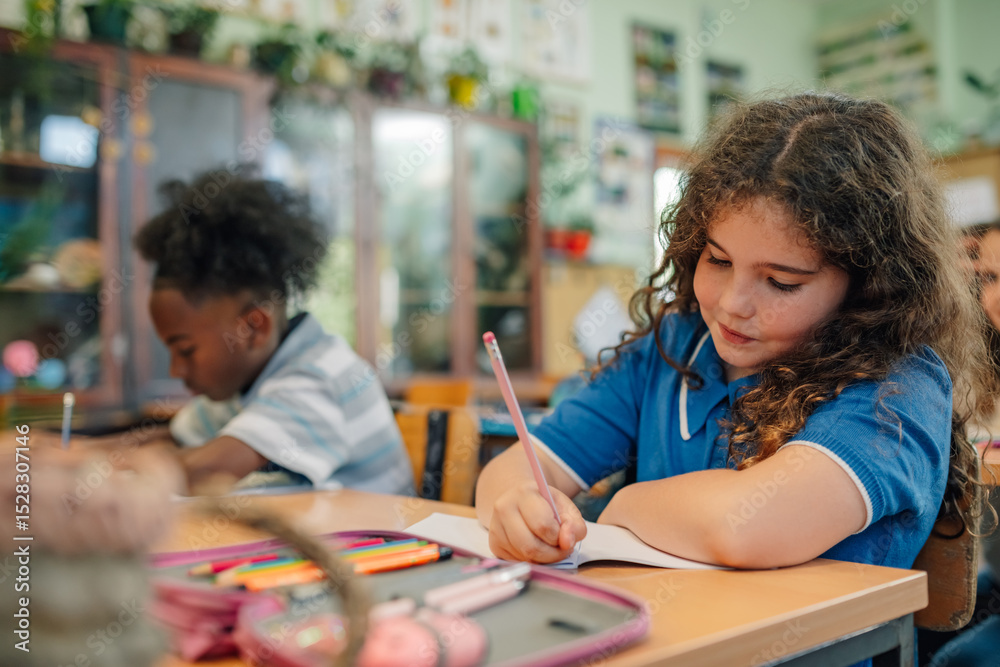 Fototapeta premium Elementary schoolgirl writing on notebook in classroom