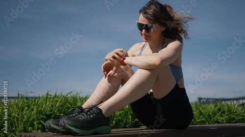 Young athletic woman wearing sportswear and sunglasses sits on a bench, checking her smartwatch after workout, with green grass and blue sky in the background