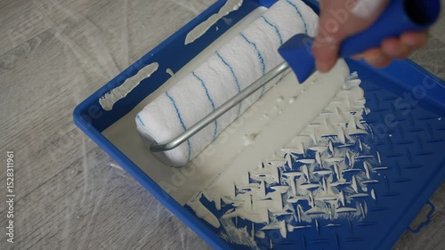 Construction worker carefully dipping paint roller in blue tray, preparing white paint for wall painting during interior home renovation project