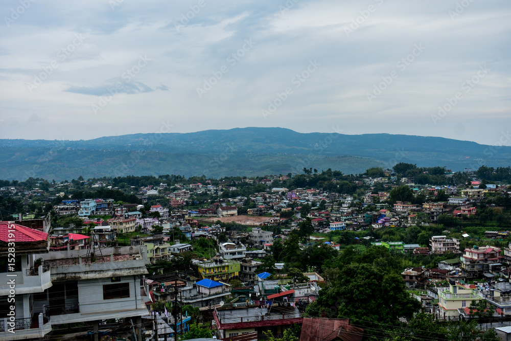 Fototapeta premium Panoramic view of a bustling hillside town near Shillong with diverse architecture under a cloudy sky.