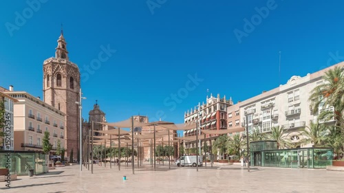 Panorama showing timelapse of Plaza de la Reina in Valencia, Spain. Lively square with a historic church tower, bustling cafes and a central fountain under the warm Mediterranean blue sky.
