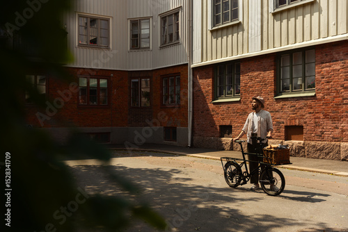 Caucasian man standing in city with cargo bicycle on a sunny day wearing a hat.