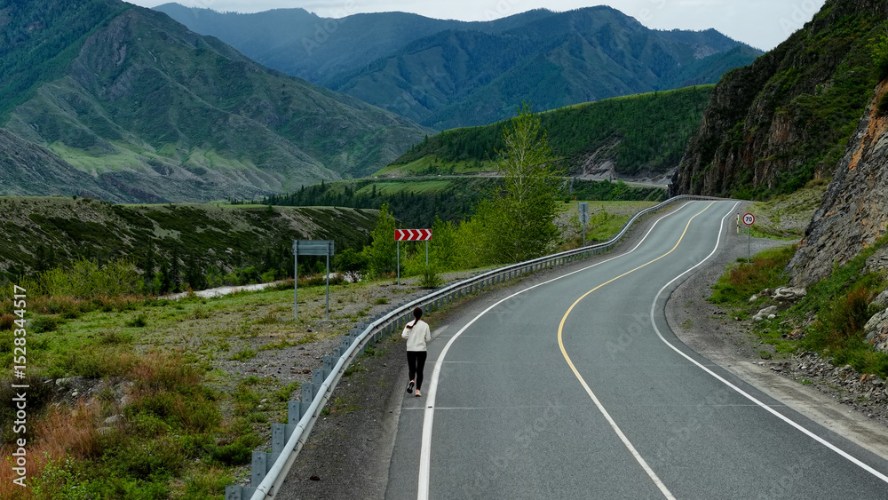 Fototapeta premium Runner in the mountains. A woman runs along a mountain road, high mountains in the background. Tourism in the Altai Mountains. Fitness while traveling.