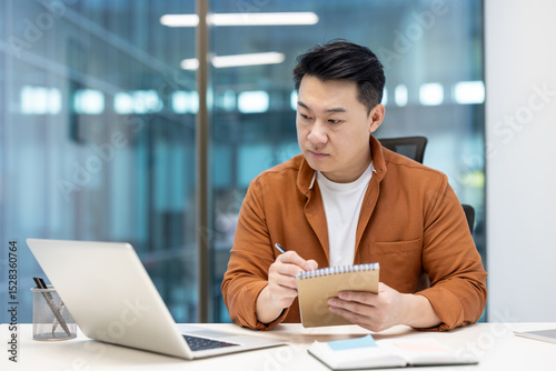 An Asian man sits at his desk, concentrating while taking notes and using his laptop in a modern office setting.
