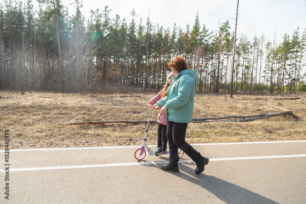 Fototapeta premium Little girl riding scooter while walking with her grandmother in the forest on a sunny day