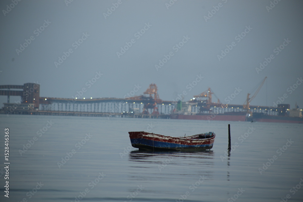 Naklejka premium Small, weathered rowboat sitting in calm water, showing signs of age and wear. In the background, a large industrial port.