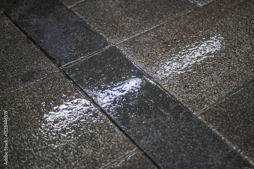 Fototapeta Naklejka Na Ścianę i Meble -  Wet stone pavement after rain. A textured surface with a mix of smooth and rough tiles, some with small puddles and water droplets.
